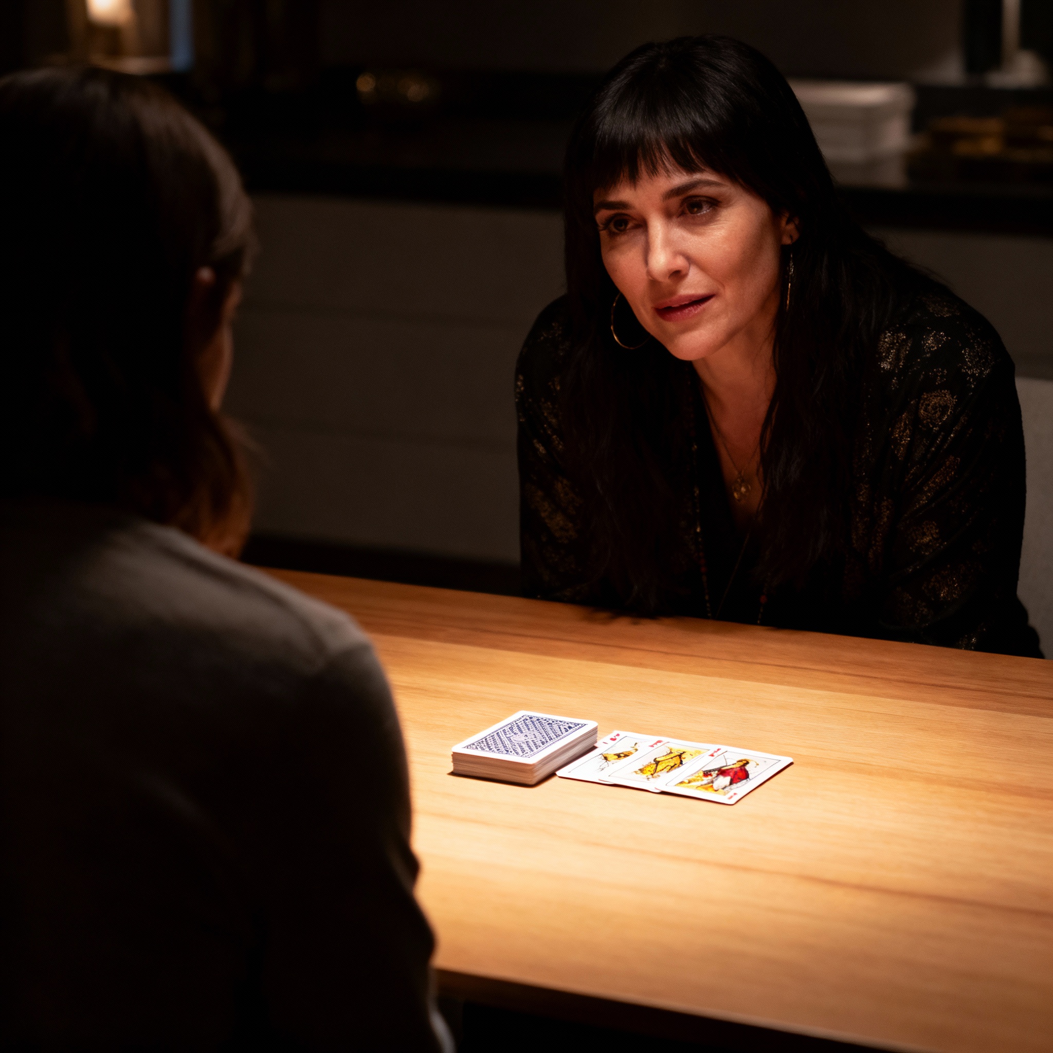During a consultation, psychic J the Oracle is speaking with a client, who is seen from behind. The focus is on the psychic's intent and empathetic expression and the face-down cards on the table, symbolizing the process of finding clarity amidst confusion.